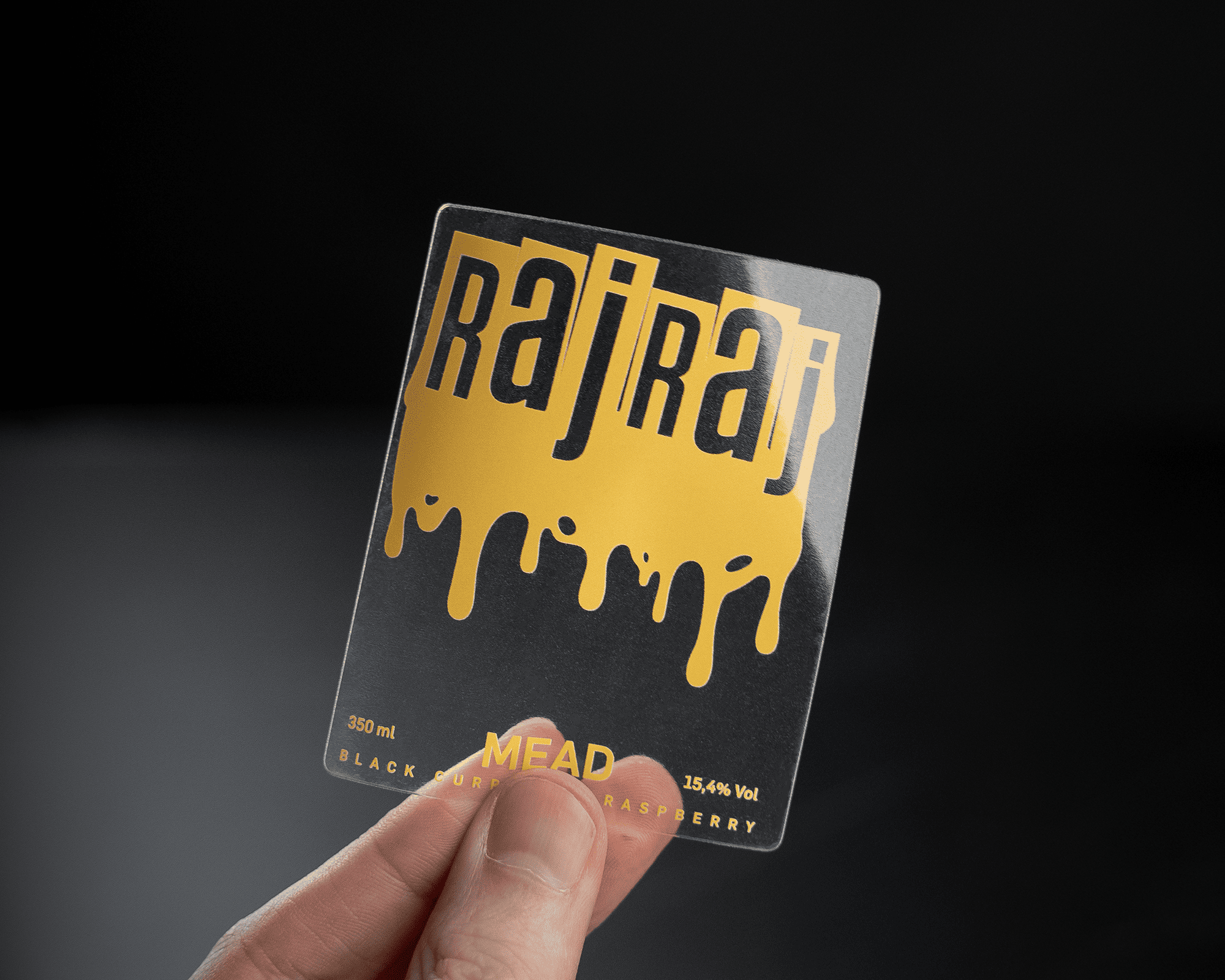 Hand holding a clear label with "Raj Raj" in bold letters and dripping gold effect, featuring details about black currant raspberry mead.